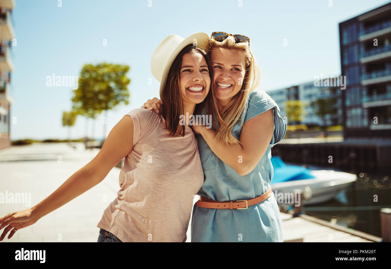 Two laughing young friends standing arm in arm together while having fun in the city in summer Stock Photo