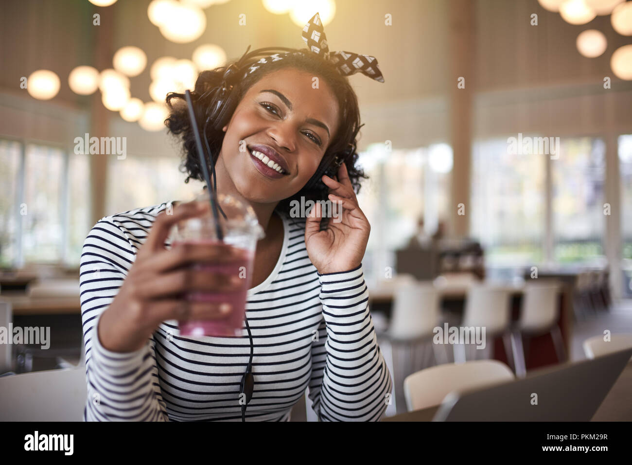 Smiling young African university student sitting on campus drinking a smoothie and listening to music on headphones while working on a laptop Stock Photo