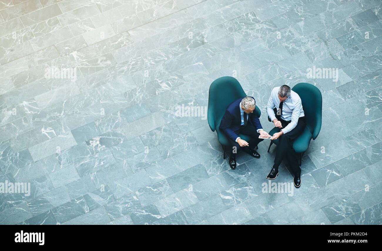 High angle of two mature businessmen sitting in the lobby of an office building talking together and using a digital tablet Stock Photo