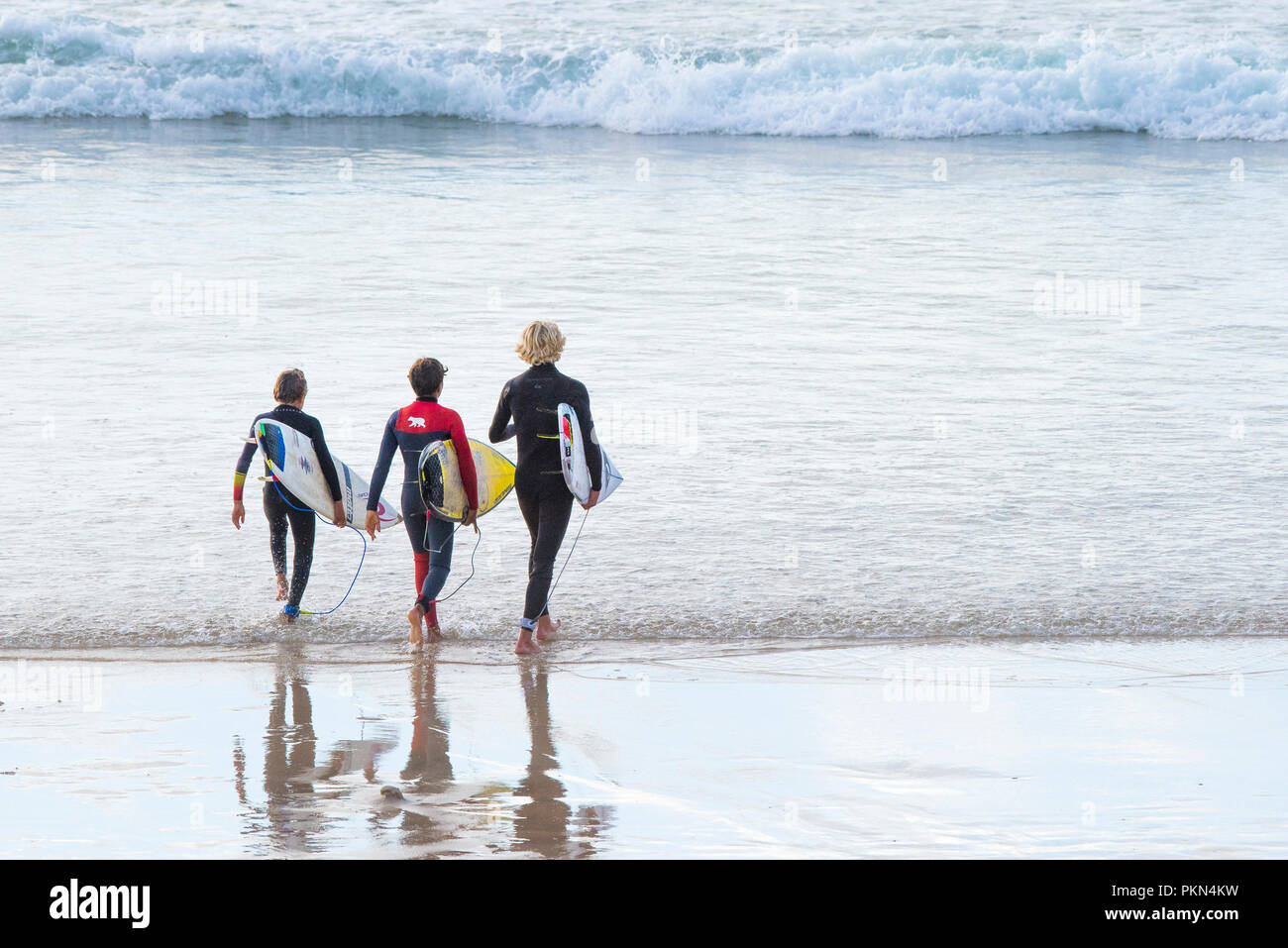 Three young surfers carrying their surfboards and walking into the sea at Fistral Beach in Newquay Cornwall. Stock Photo