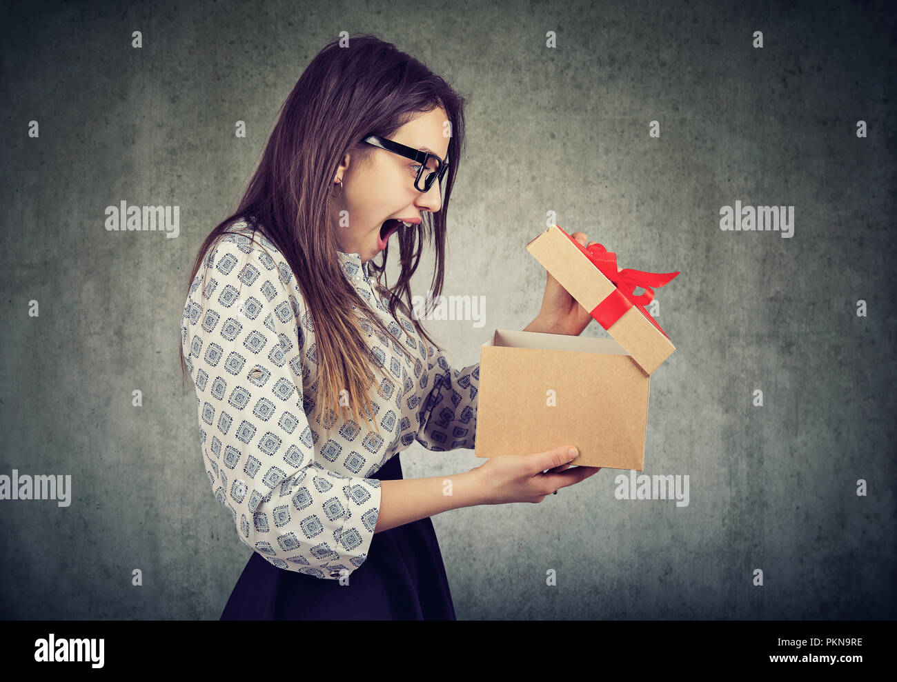 Side view of casual woman in glasses opening present box with bow looking super amazed on gray background Stock Photo
