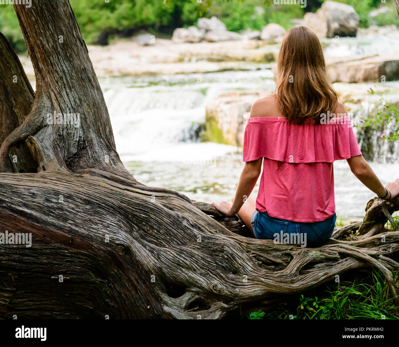 Teen Girl With Long Brown Hair Sitting On A Large Tree Branch With Waterfall In The Background Stock Photo