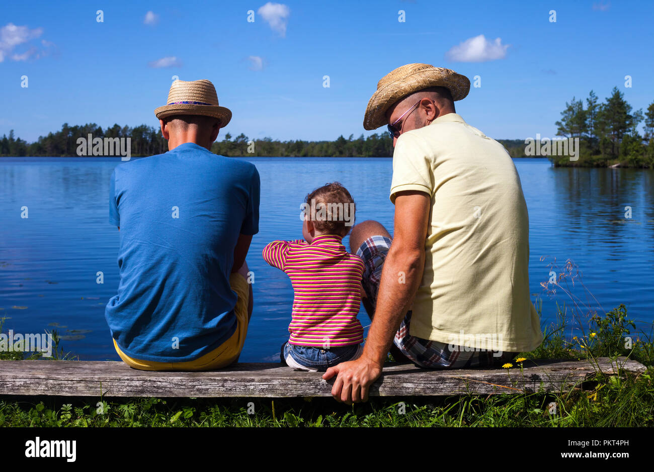 Redhead toddler sitting on a bench near a river with two men on the sides - gay parenting concept Stock Photo