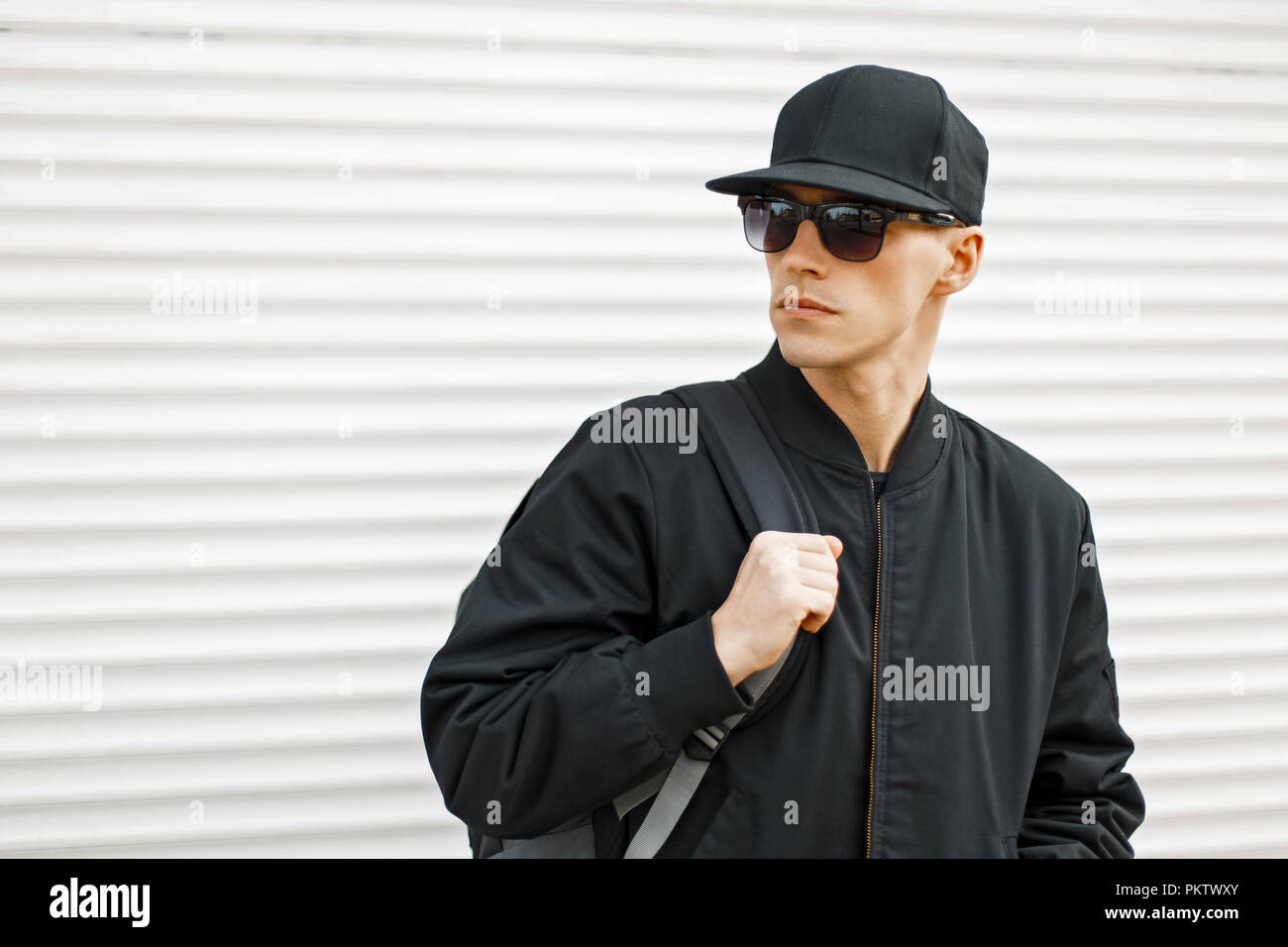 Handsome young man with sunglasses in a black baseball cap and jacket with  a bag Stock Photo - Alamy