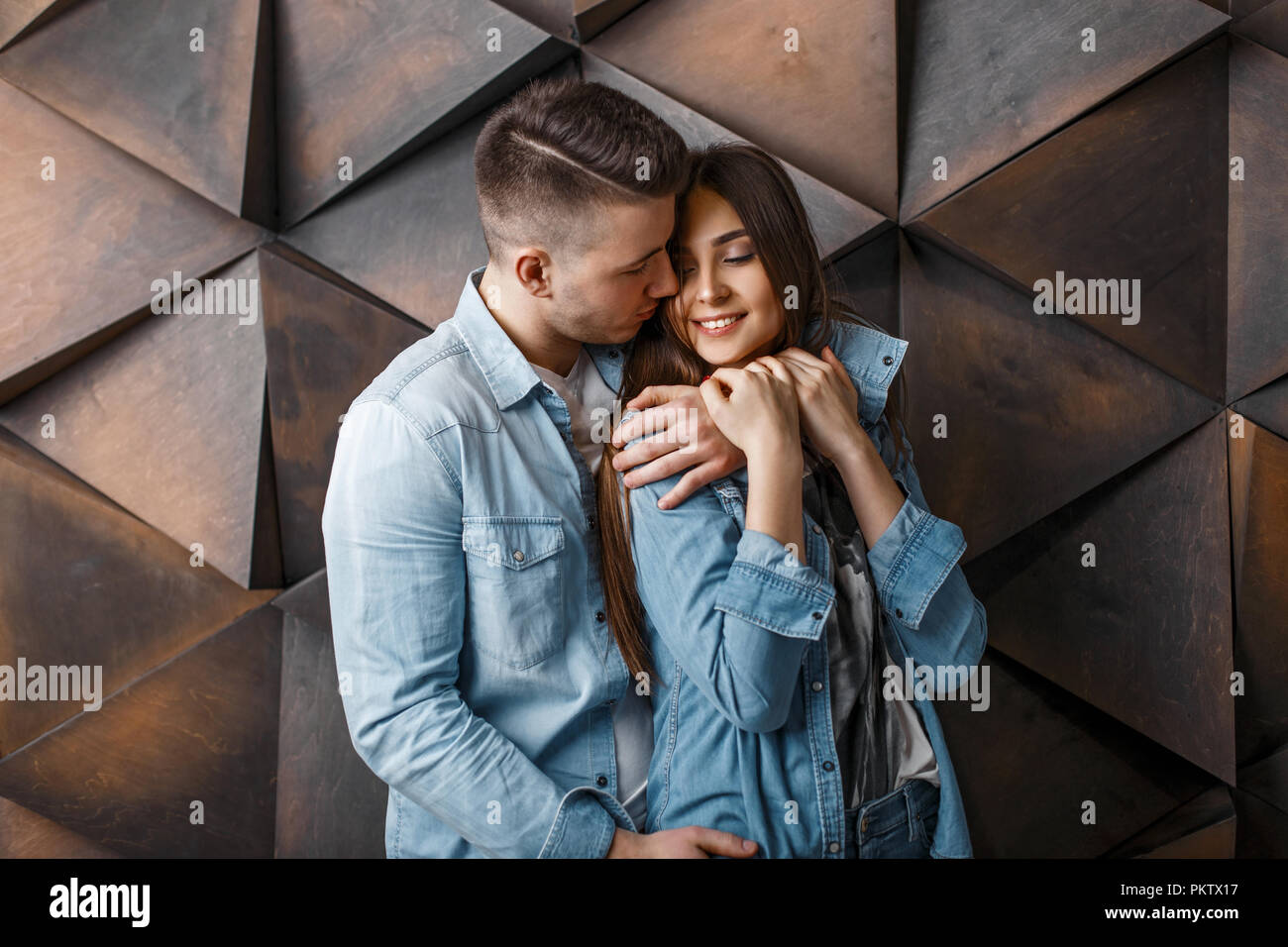 Beautiful happy young couple in blue jeans clothes hug near a wooden patterned wall Stock Photo