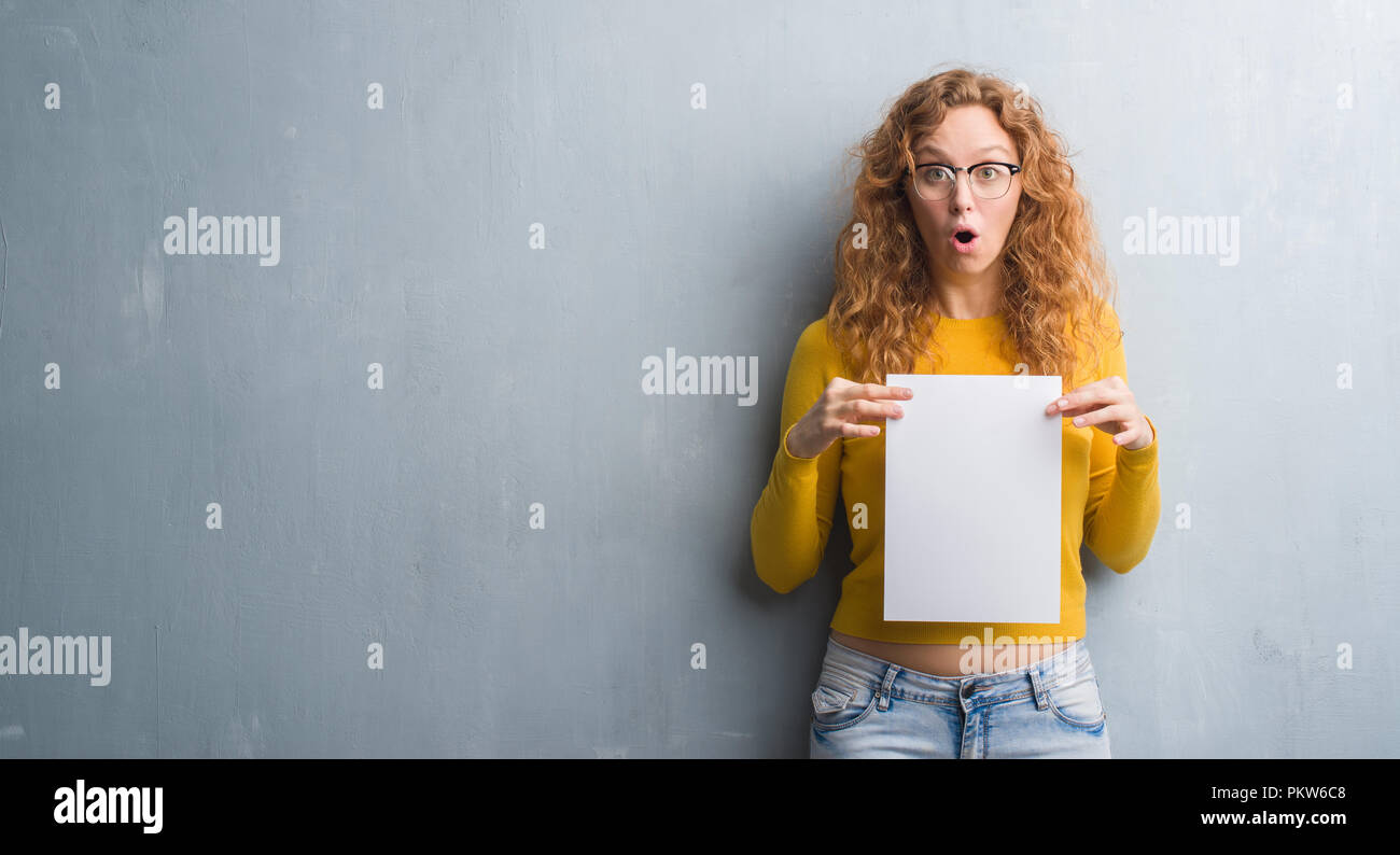 Afraid Paper Young redhead woman over grey grunge wall holding blank paper sheet ...