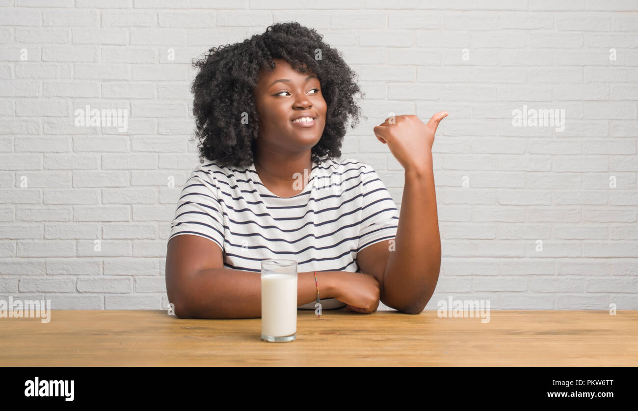 Young african american woman sitting on the table drinking a glass of milk pointing and showing with thumb up to the side with happy face smiling Stock Photo