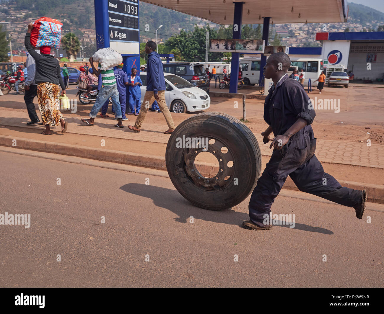 a man in overalls running past a fuel station with a truck tyre/wheel on the road in Kigali, rwanda Stock Photo