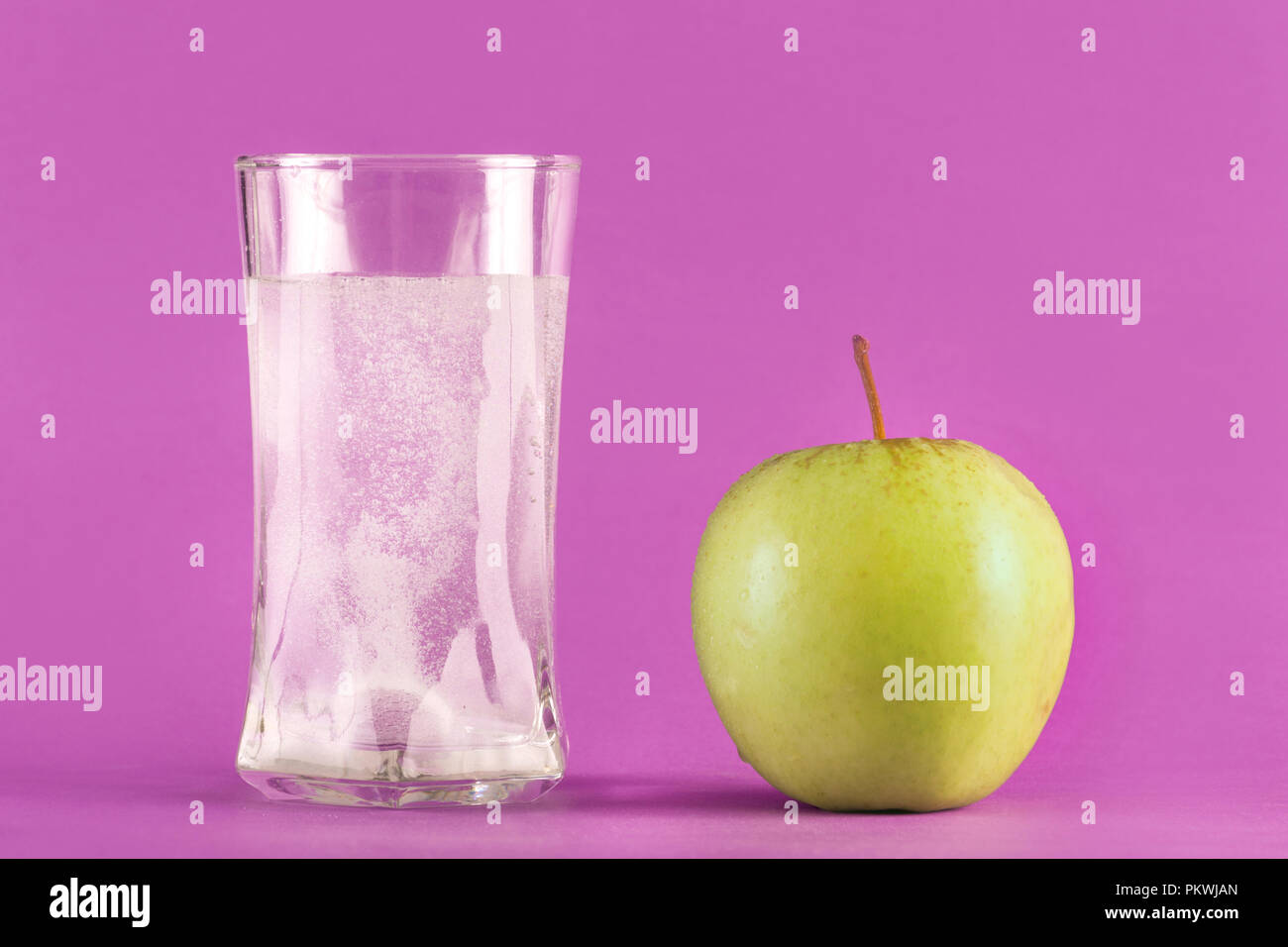 Water glass with effervescent tablet with bubbles and green apple isolated on purple background. Medical and health concept Stock Photo