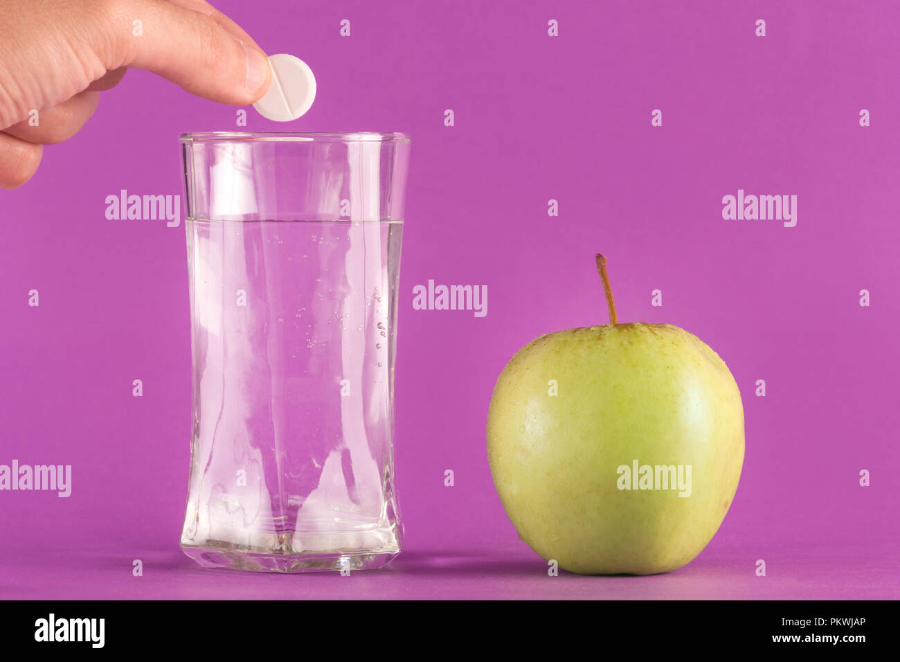 Hand throwing effervescent tablet vitamin mineral supplement into glass of water and green apple isolated on purple background. Health and medical Stock Photo