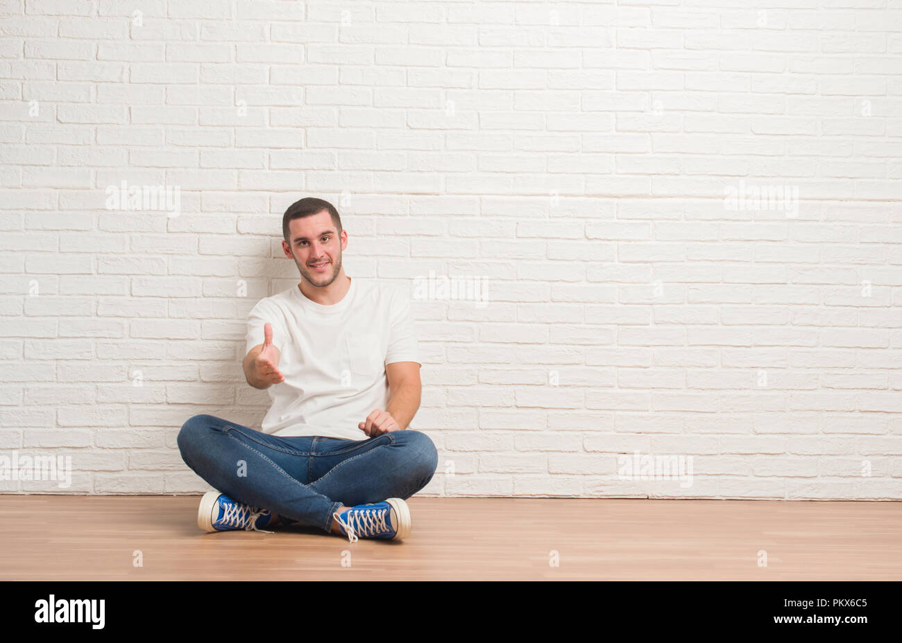 Young caucasian man sitting on the floor over white brick wall smiling