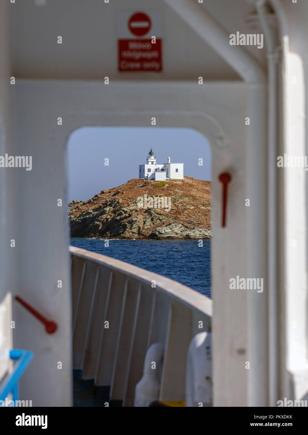 View from ship's door of lighthouse and agios Nikolaos church on rocky land. Kea,Tzia island, Greece. Blue sky background. Stock Photo