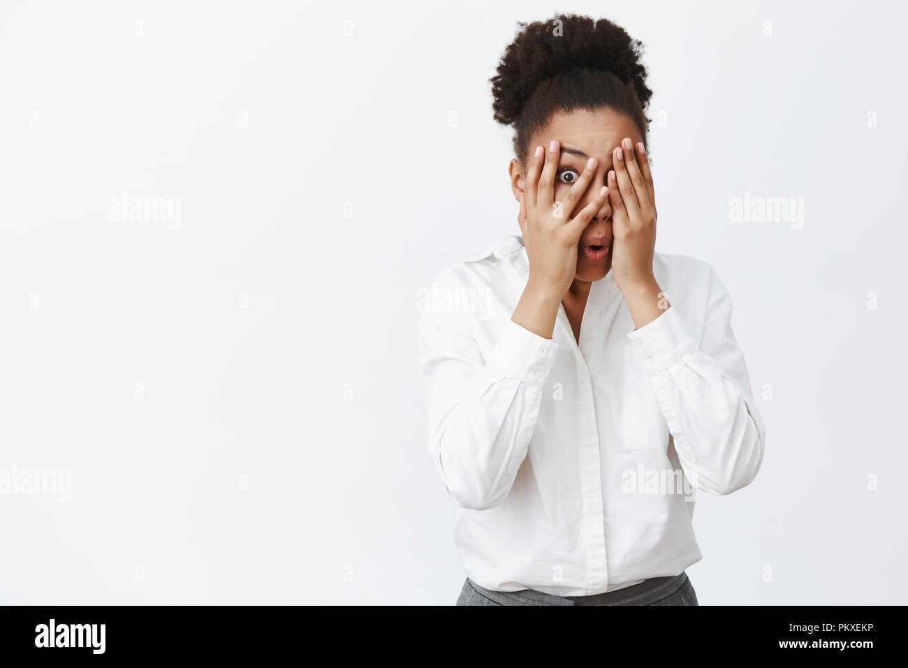 Woman feeling scared but curious, peeking through fingers with one eye while covering sight with palms, folding lips from shock and amazement, standing frightened over gray background Stock Photo