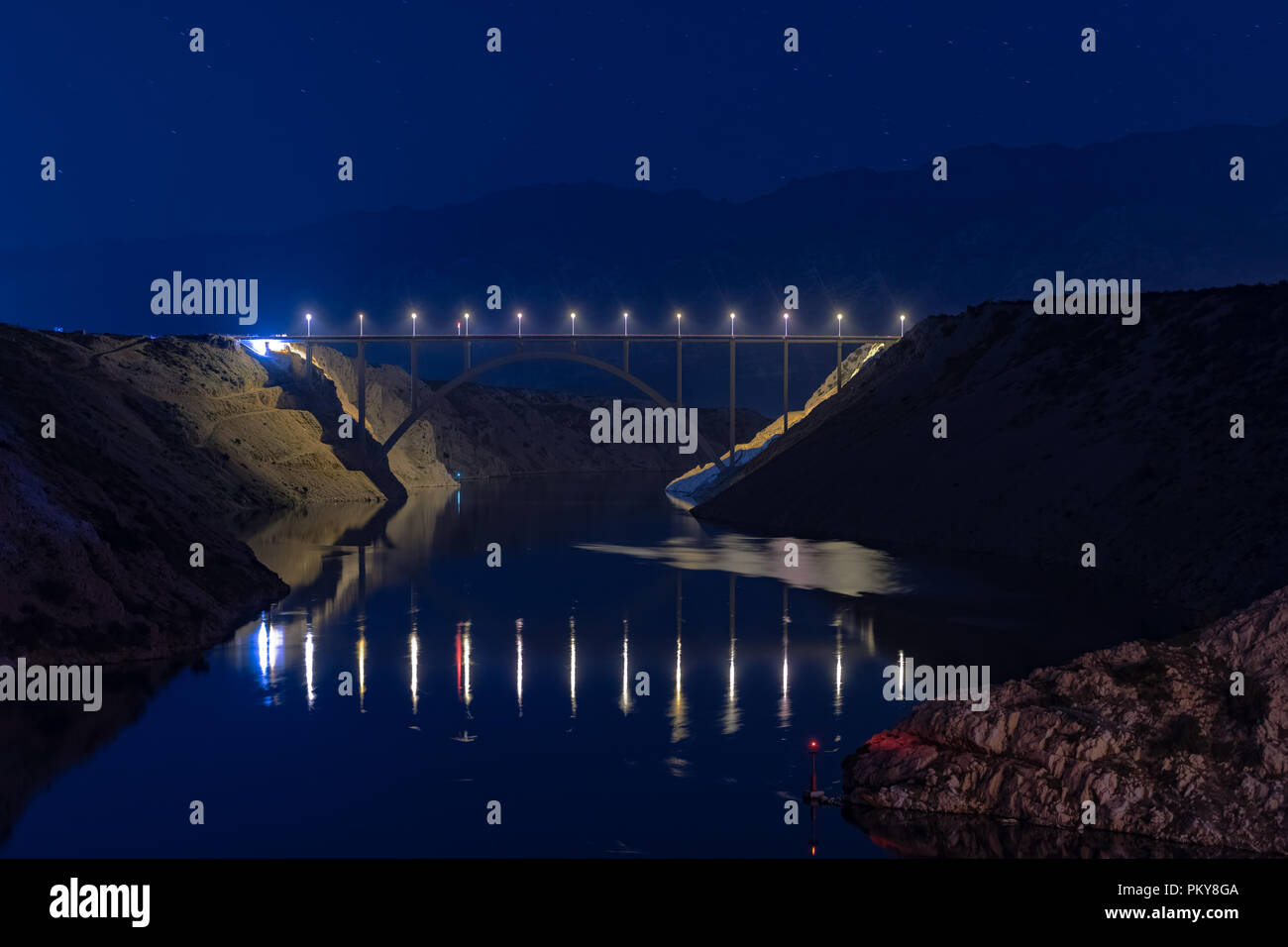 Illuminated highway bridge under stars, reflection of lights in water, mountains in background and bright stars in sky Stock Photo