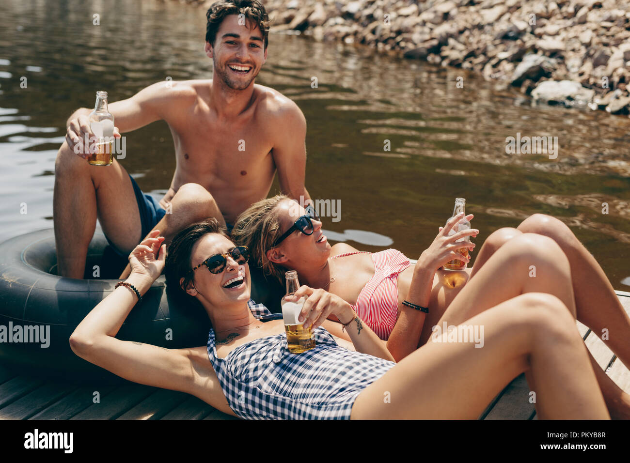 Man with two woman friends sitting on a floating dock in a lake drinking beer. Friends having fun at a lake relaxing with drinks in hand. Stock Photo