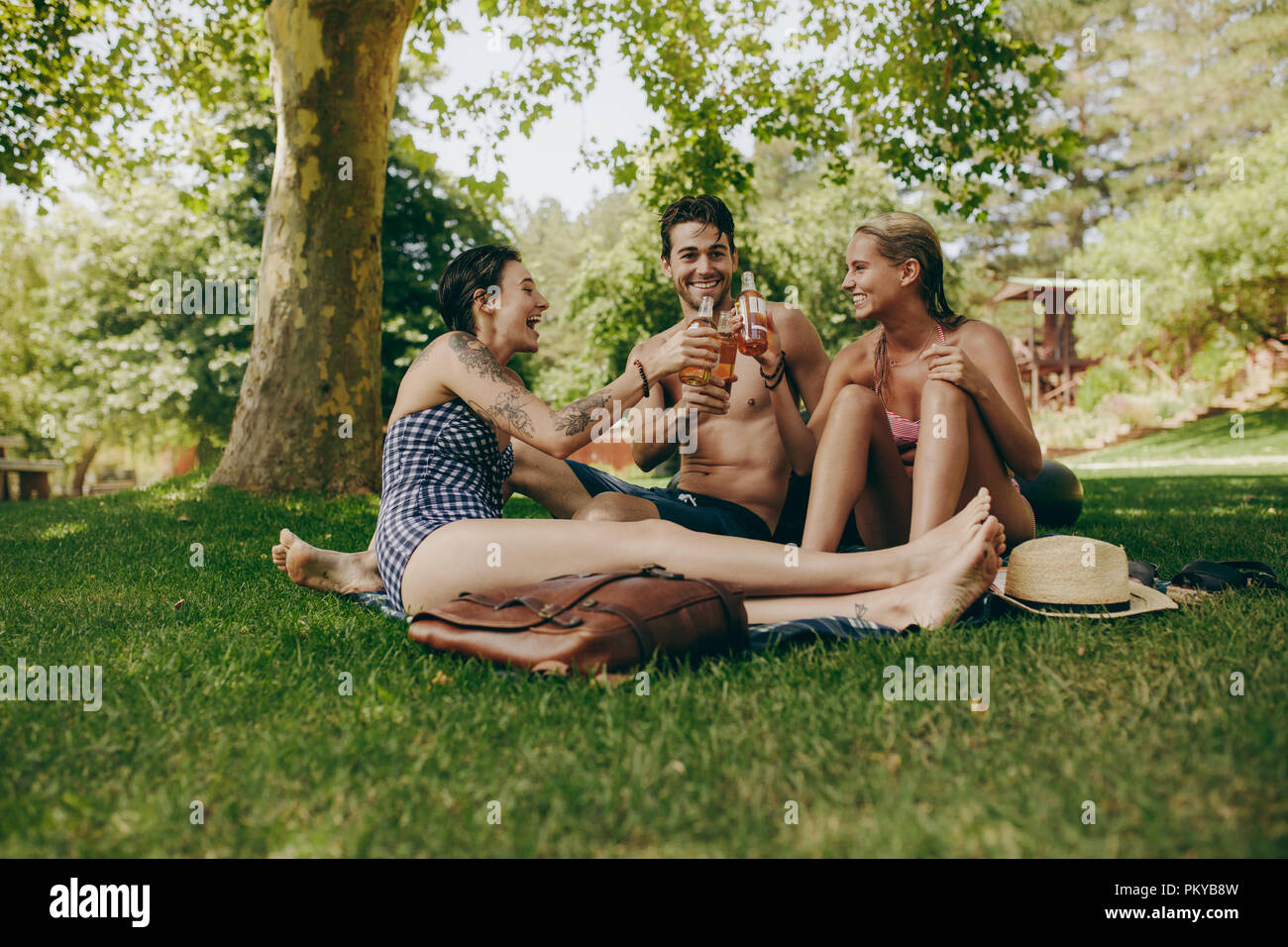 Two women in bikini toasting their drinks with a male friend sitting in a park. Friends enjoying and relaxing in park with drinks in hand. Stock Photo
