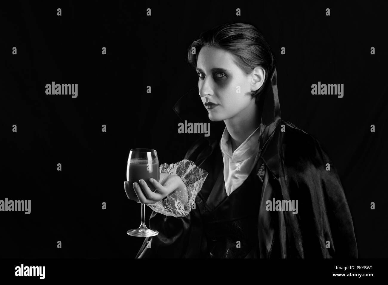 Woman with a glass of red drink in vampire costume. Young female with glass of red drink dressed up as dracula posing in studio. Stock Photo