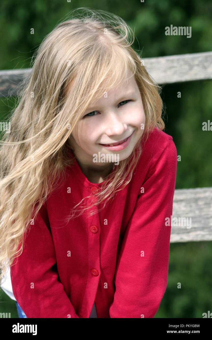 little blonde girl with curly long hair, portrait, in a red sweater in the sun, vertical photo, looks at the camera and smiles, in the background Stock Photo