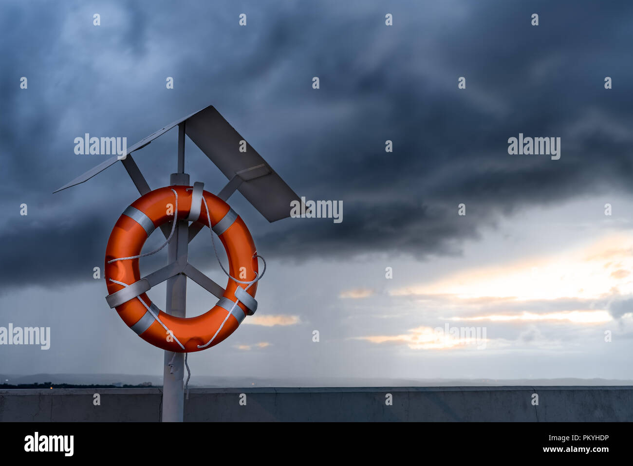 Orange lifebuoy on a stand on harbor pier against dramatic clouds in the sky. Stock Photo