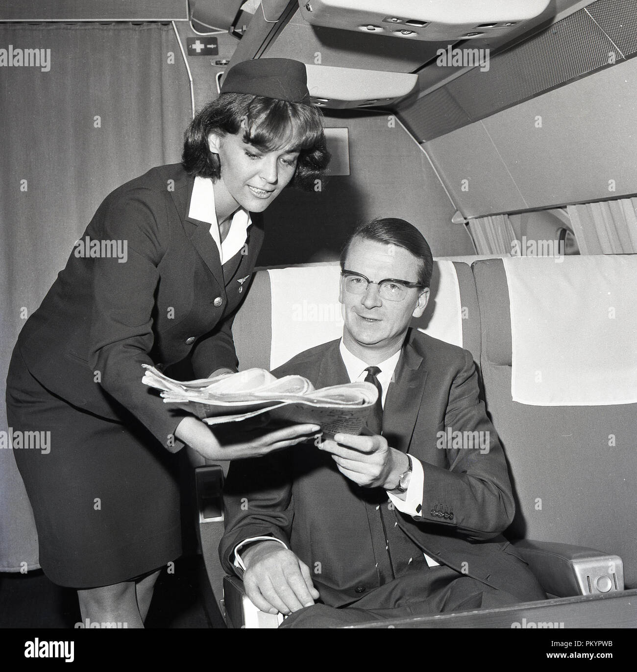 1960s, historical, a uniformed female flight attendant presenting a daily newspaper to a suited male passenger sitting in a business class seat on an aircraft. Stock Photo