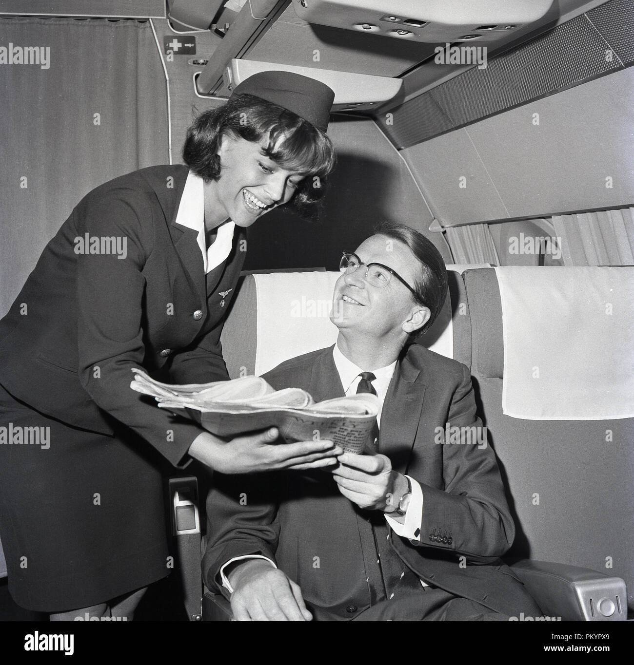 1960s, historical, a uniformed female flight attendant presenting a daily newspaper to a male passenger sitting in a business class seat on the aircraft. Stock Photo
