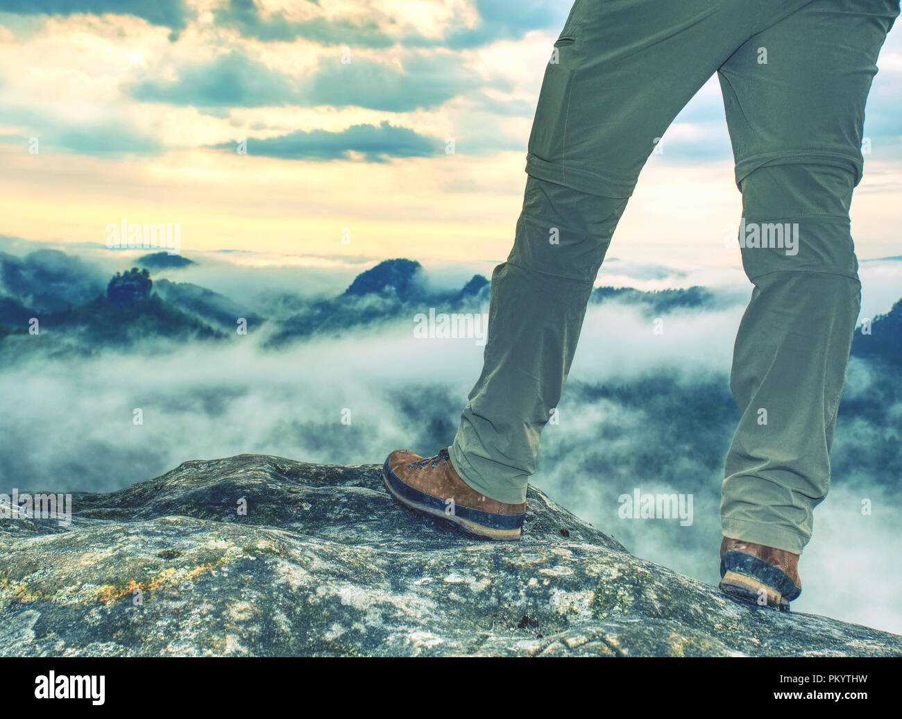 Legs in hiking trousers and leather trekking shoes on peak of rock above misty valley. Autumnal nature  Outline of hill side on horizon Sun hidden in  Stock Photo