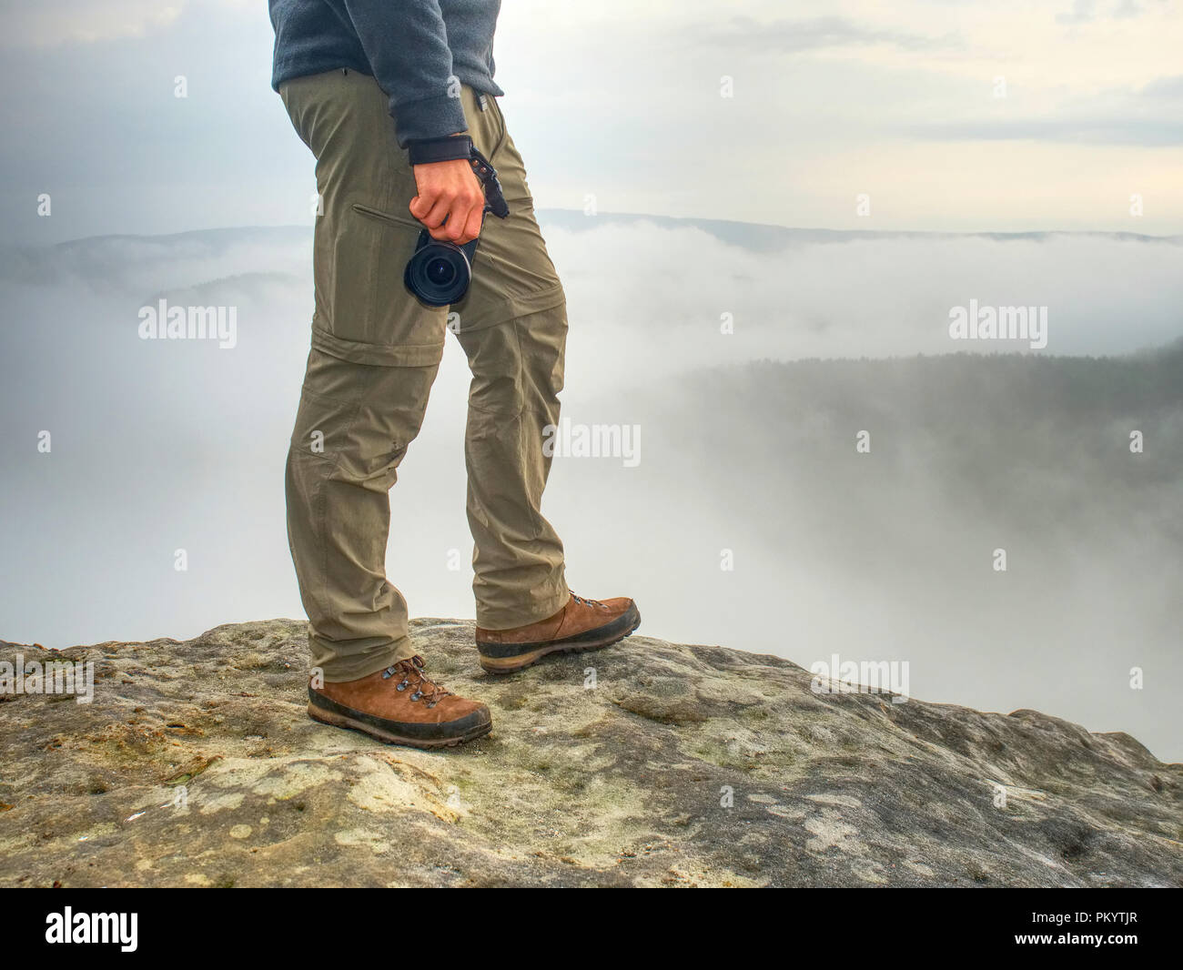 Photographer hand with  camera,  legs, knees and boots. Man takes landscape photos on peak of rock. Dreamy fall  landscape Sun colorized horizon Stock Photo