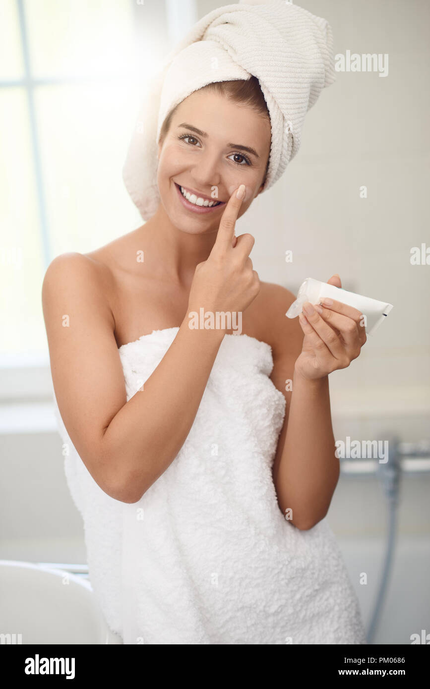 Pretty young woman in white towels in a bathroom giving the camera a lovely friendly smile as she applies cream to her cheeks while bathing in a skin  Stock Photo