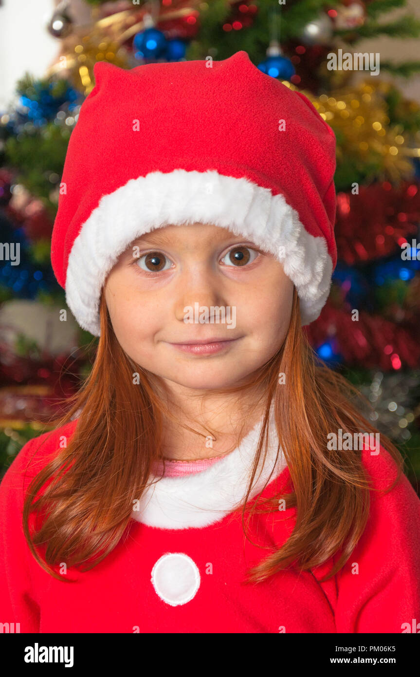 little redhair girl dressed as Santa Claus. smiling, red cup on head. colorful christmas tree with decorations in background Stock Photo