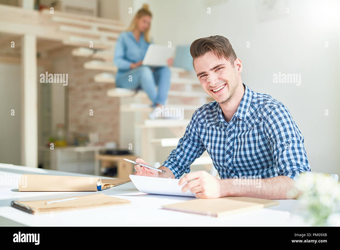 Laughing young designer sitting by table and reading papers while preparing for new project Stock Photo