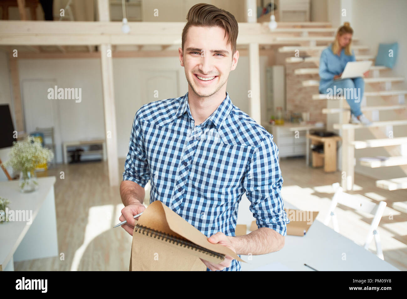 Smiling young casual man with notepad making working notes while working in studio on sunny day Stock Photo