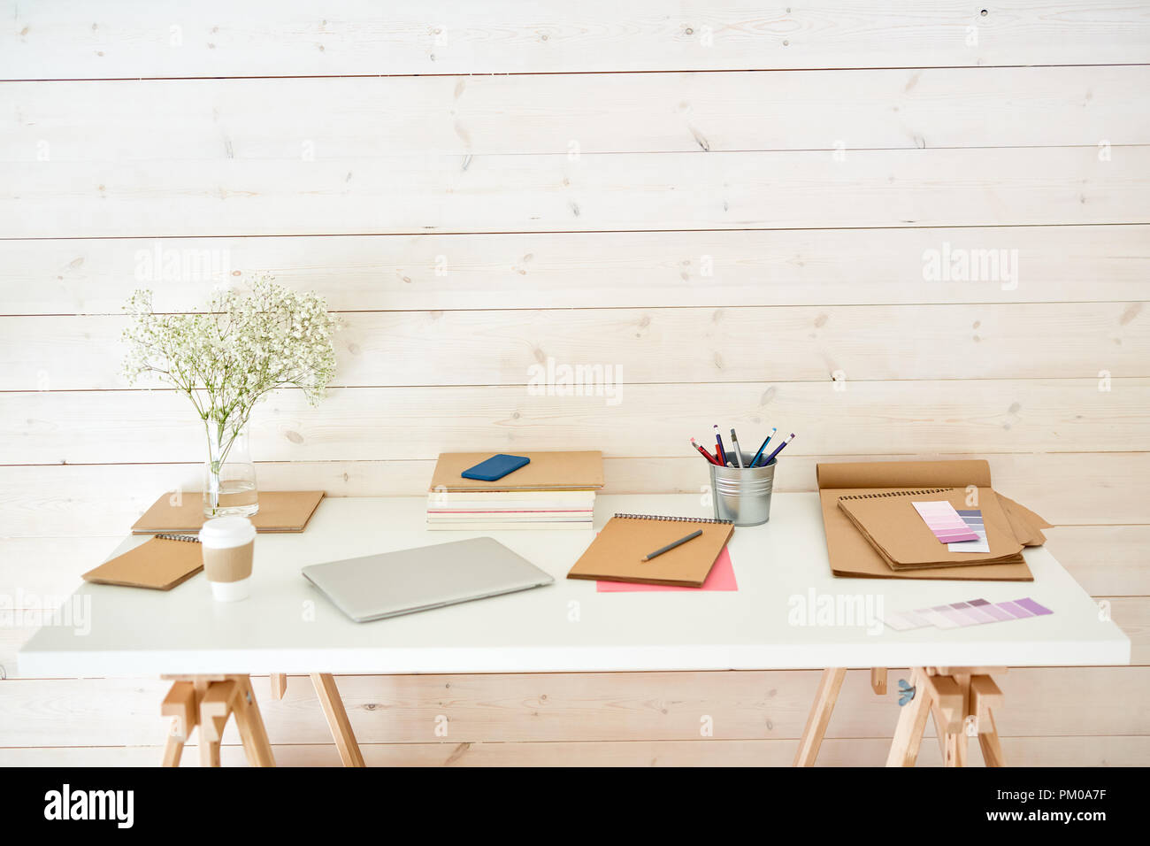 White desk by wall of studio with drink, gadgets, notepads, crayons in small bucket and some