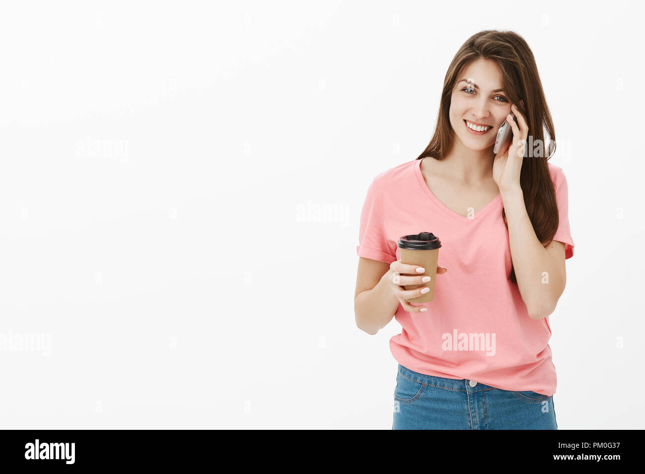 Portrait of joyfull good-looking girlfriend in trendy urban outfit talking on smartphone casually and smiling broadly at camera while talking with friend and drinking coffee, walking to office Stock Photo