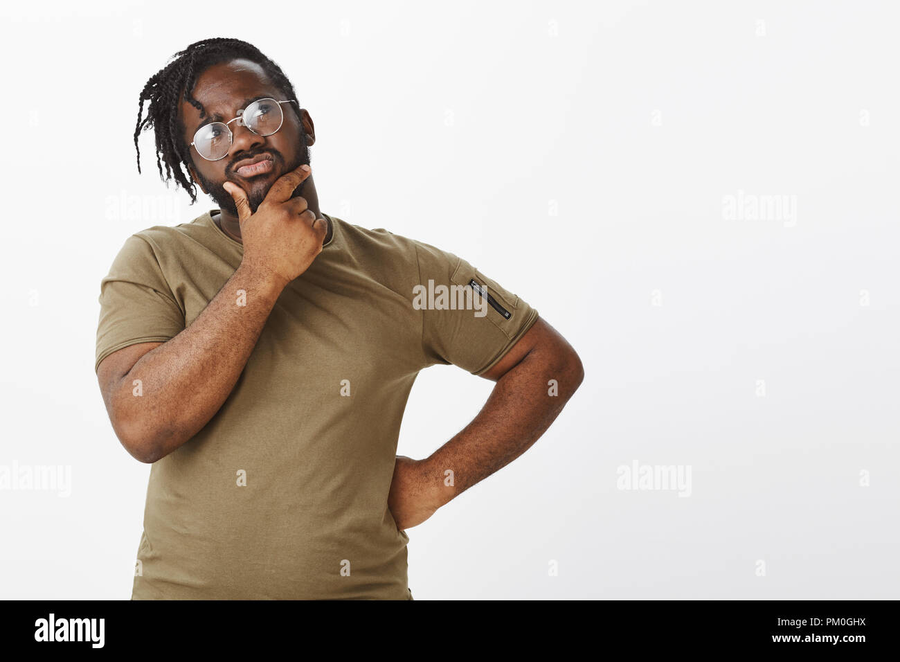 Hmm, what should guy do. Portrait of troubled focused handsome man with dark skin in t-shirt and glasses, holding hand on hip and rubbing chin while looking thoughtfully at upper right corner Stock Photo