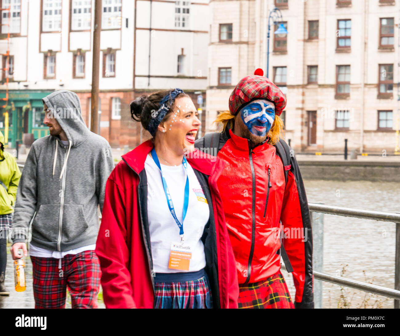 Leith, Edinburgh, Scotland, UK, 16th September 2018. Edinburgh Kilt Walk, sponsored by the Royal Bank of Scotland, takes place today. Walkers raise funds for a charity of their choice. The kilt walkers reach The Shore at about Mile 14 in the rain. A couple laughing as they walk on the riverbank. The man is wearing a See you Jimmy tartan hat and has a saltire painted on his face Stock Photo