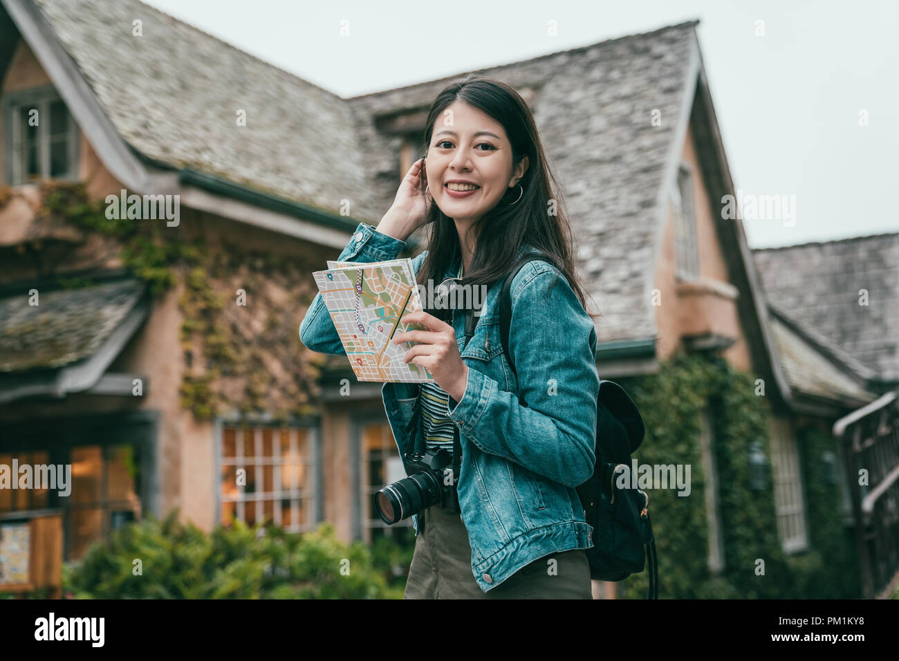 woman smiling joyfully and holding a guide map in hands while walking into a small and stunning town. Stock Photo