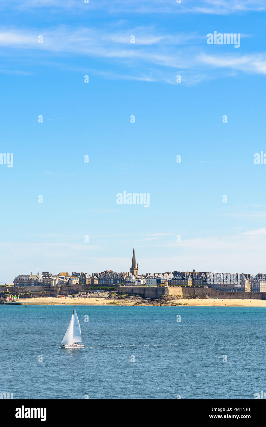 The walled city of Saint-Malo in Brittany, France, with the cathedral protruding above the buildings behind the wall and a boat sailing on the sea. Stock Photo