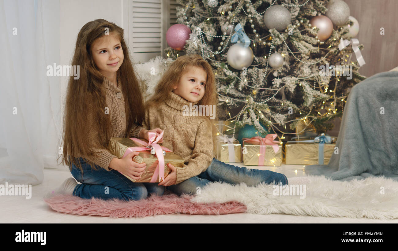 Two little sisters are waiting for Christmas. Girls are sitting by Christmas tree and holding gifts. They are dressed in jeans and sweaters. Happy chi Stock Photo