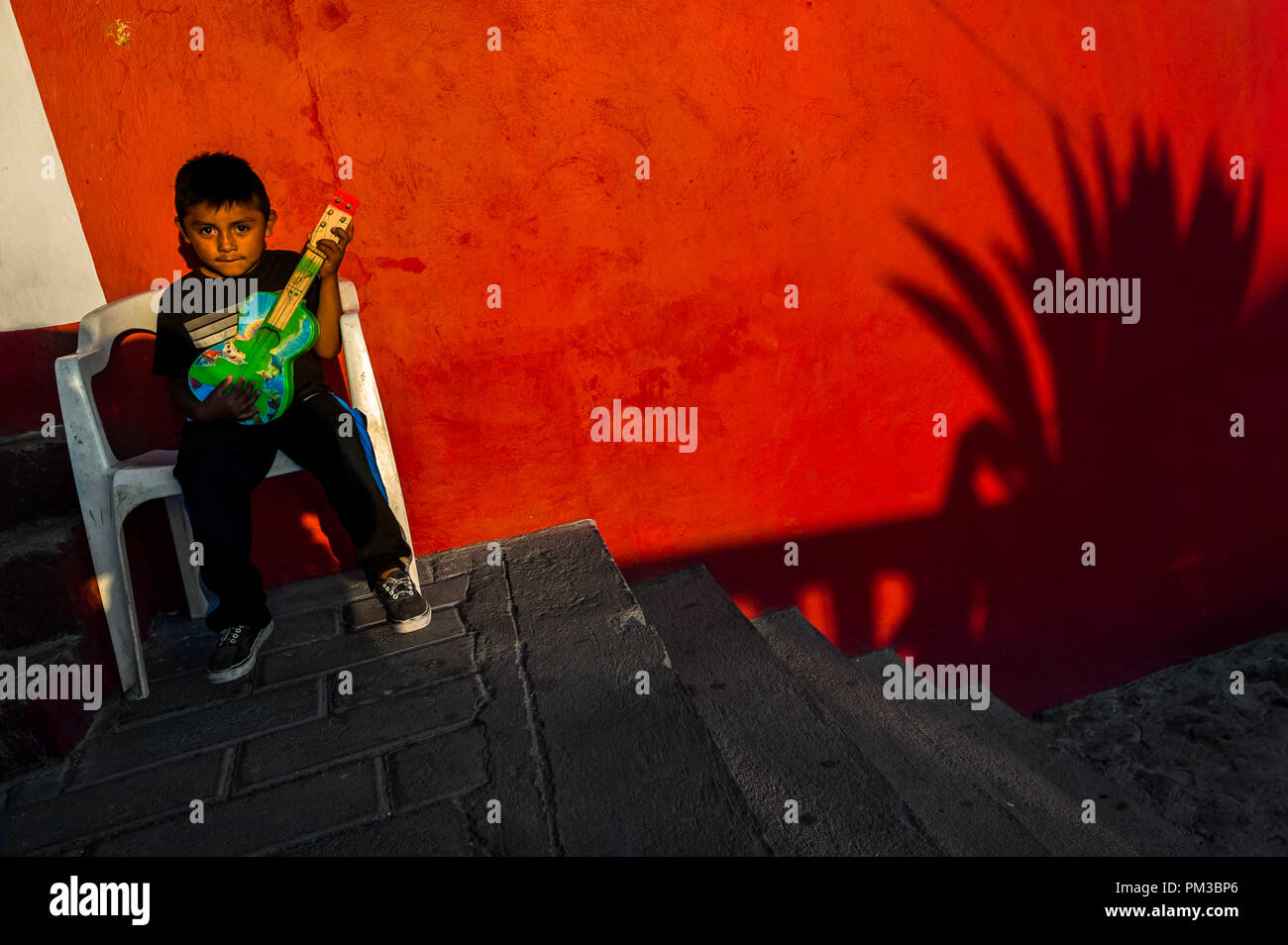 A Mexican boy plays a toy guitar while an agave succulent shadow is seen on the wall of a house in Atlixco, Mexico. Stock Photo