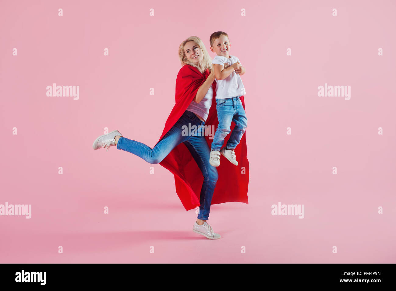 Mom is a superhero. Fun family, a young blond woman in a red Cape and her son jumps and takes off, pink background Stock Photo