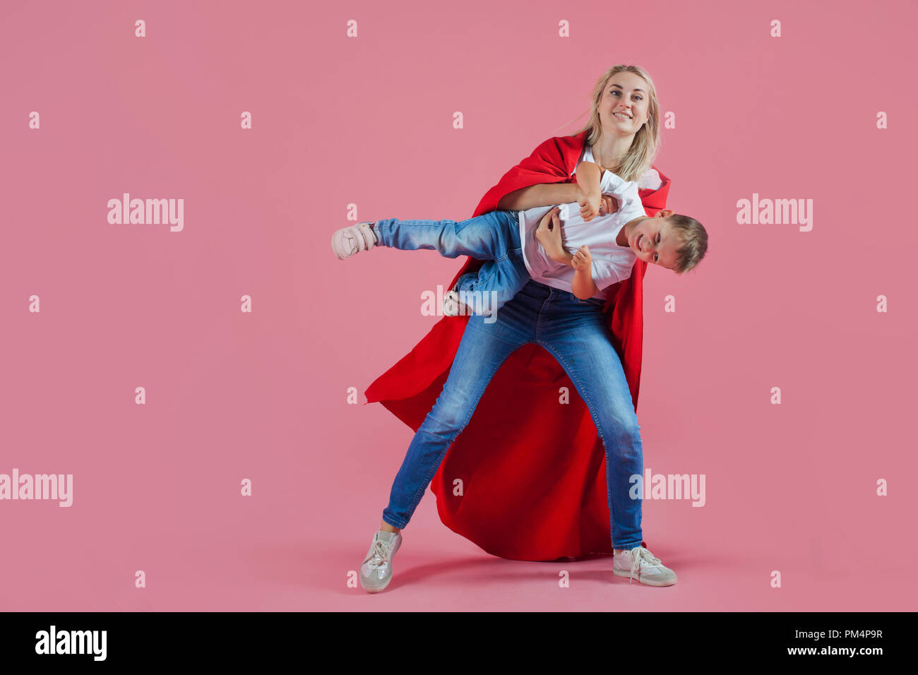 Mom is a superhero. happy family, a young blond woman in a red Cape and her son jumps and takes off, pink background Stock Photo