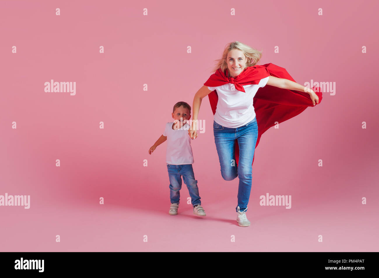 super mom and her son run forward. Fun family, a young blond woman in a red Cape as superhero, pink background Stock Photo