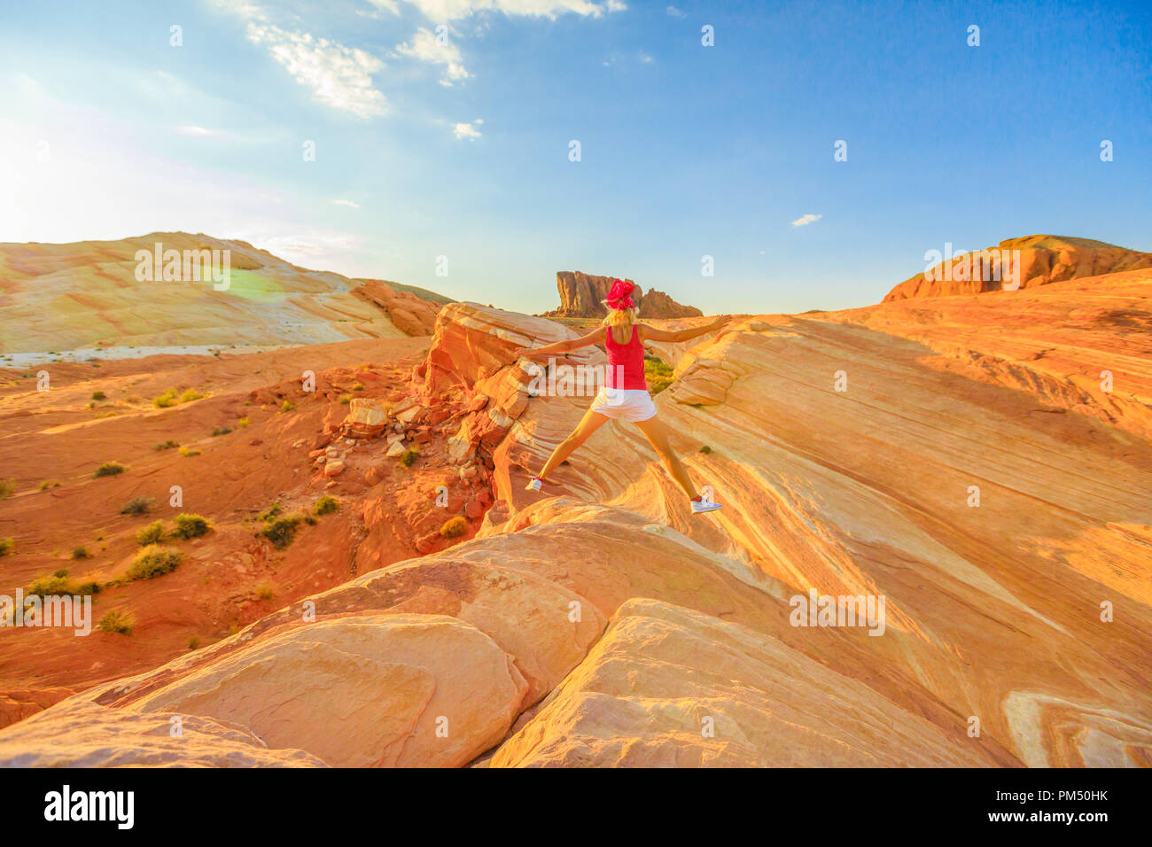 Young tourist girl jumping over Fire Wave in Valley of Fire, Nevada, USA, at red sunset. Happy hiker enjoying view of popular striped lines around the lip of Fire Wave. Gibraltar rock on background. Stock Photo