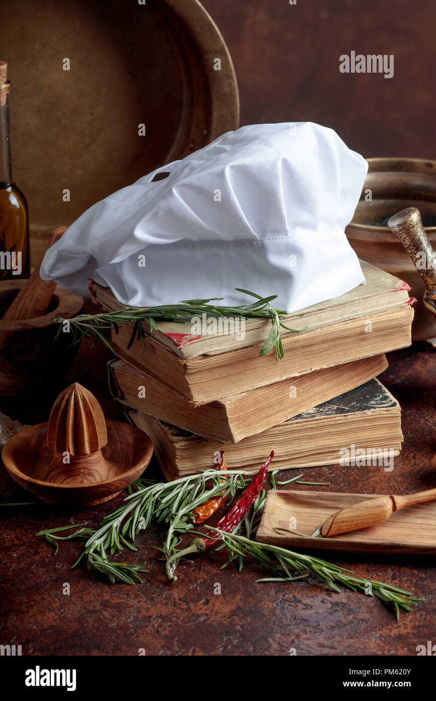 White chef's hat and old cookbooks. Kitchen utensils with spices and rosemary on a kitchen table. Stock Photo