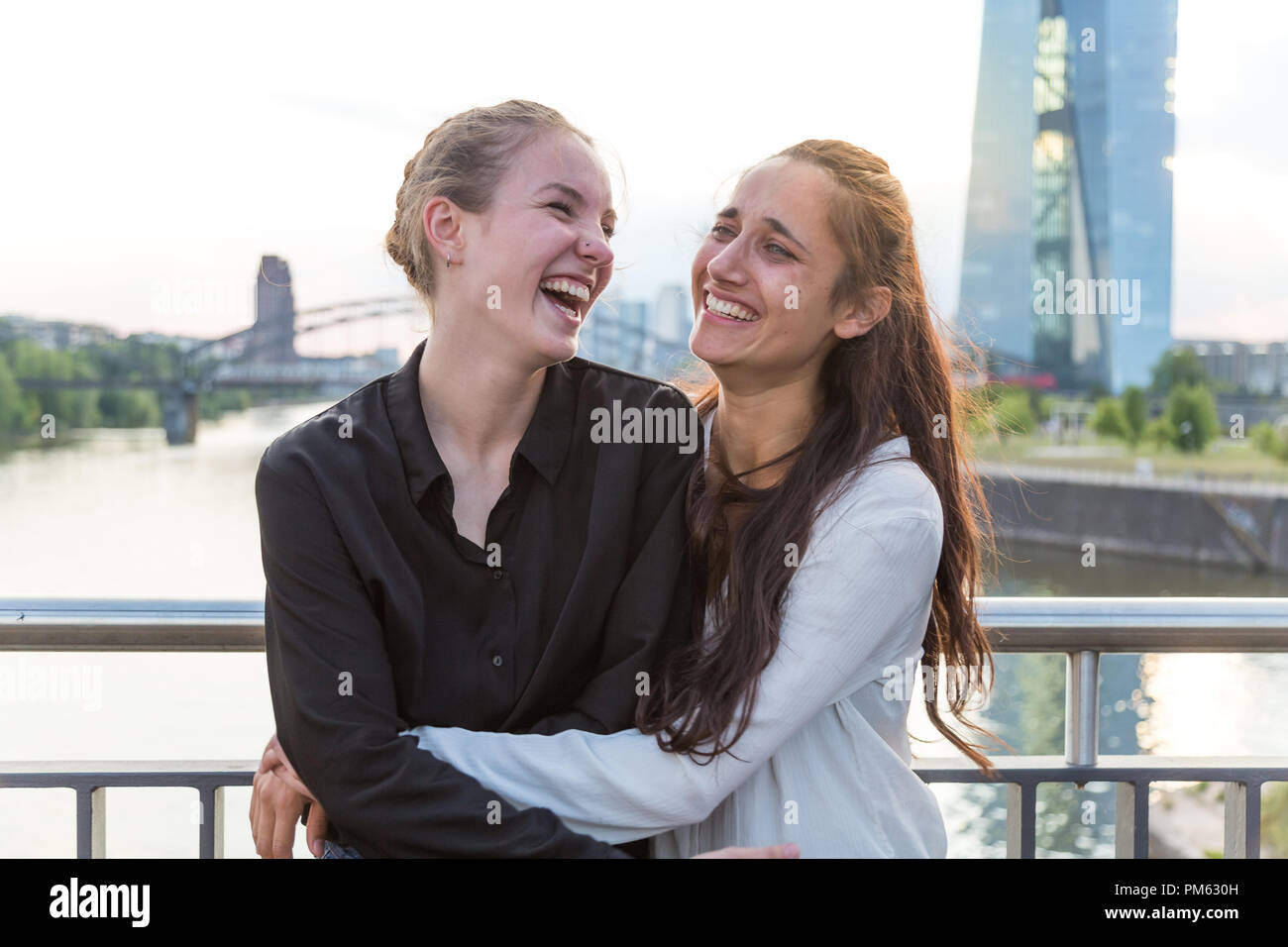 Girlfriends laughing wildly and embracing on city bridge over river with metropolis skyline in the background. Medium shot. Stock Photo