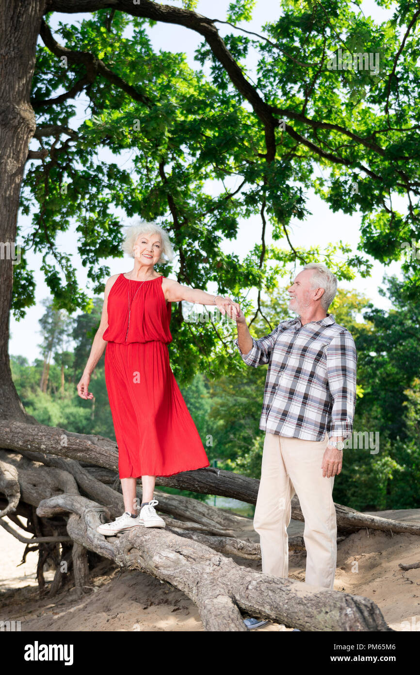 Happy pensioner holding hand of his wife and looking at her standing on the log Stock Photo