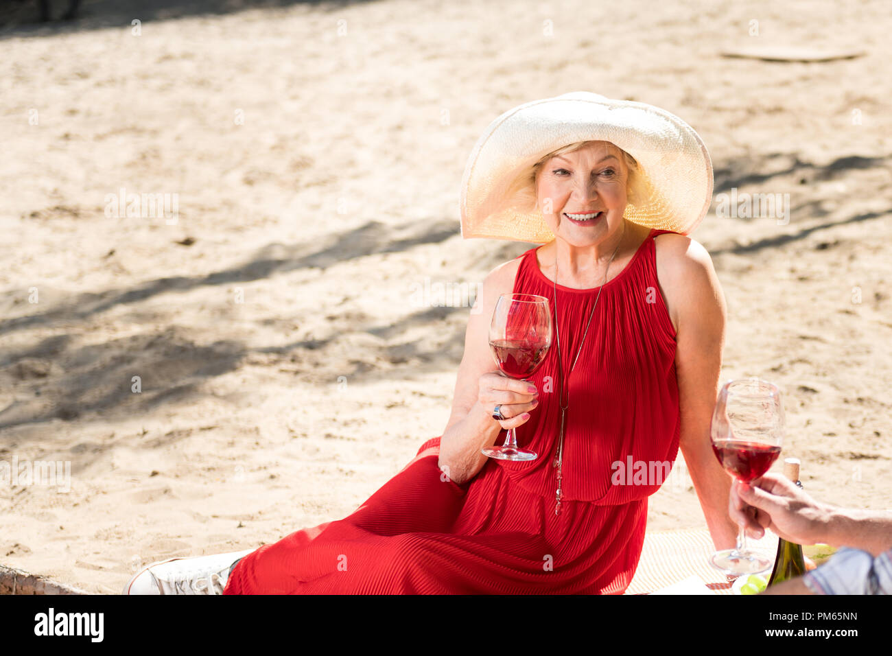 Happy aged woman sitting on the sand and drinking wine Stock Photo