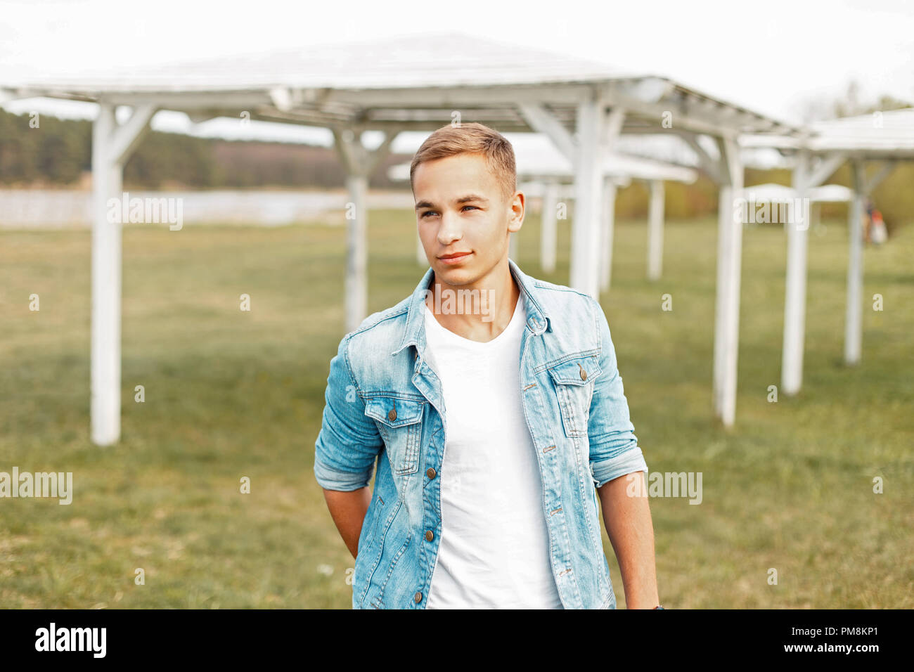 Young man in a stylish denim jacket and a white T-shirt is walking outdoors on a spring day Stock Photo