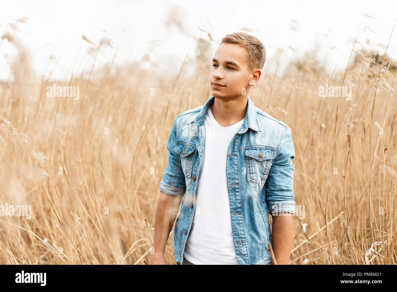 Handsome young man in a jeans jacket in a white T-shirt is walking outdoors in the grass Stock Photo