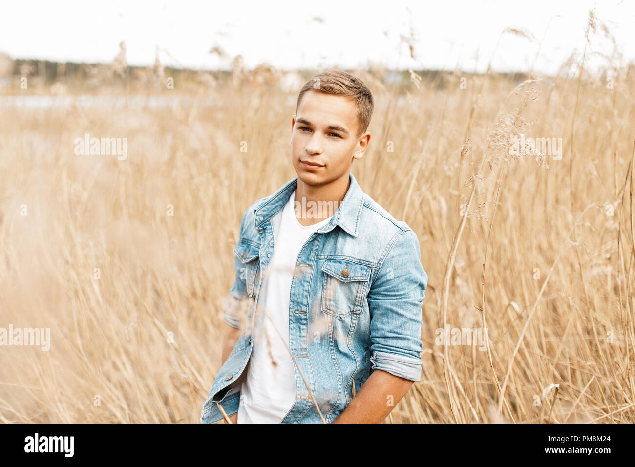 Handsome young man in a jeans jacket and a white T-shirt in dry grass walks in nature Stock Photo